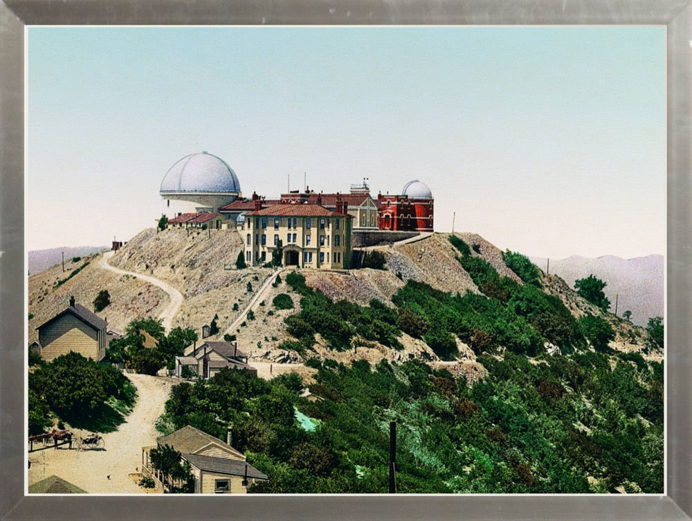 A print of Lick Observatory, located atop Mt. Hamilton in San Jose, California. The observatory's beautiful buildings are set against a mountain backdrop with lush foliage and a bluish-pink sky.