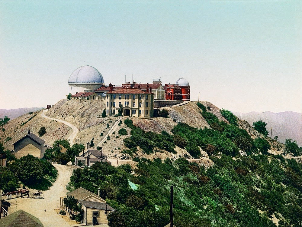 A print of Lick Observatory, located atop Mt. Hamilton in San Jose, California. The observatory's beautiful buildings are set against a mountain backdrop with lush foliage and a bluish-pink sky.