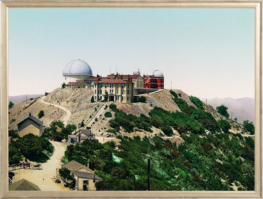 A print of Lick Observatory, located atop Mt. Hamilton in San Jose, California. The observatory's beautiful buildings are set against a mountain backdrop with lush foliage and a bluish-pink sky.